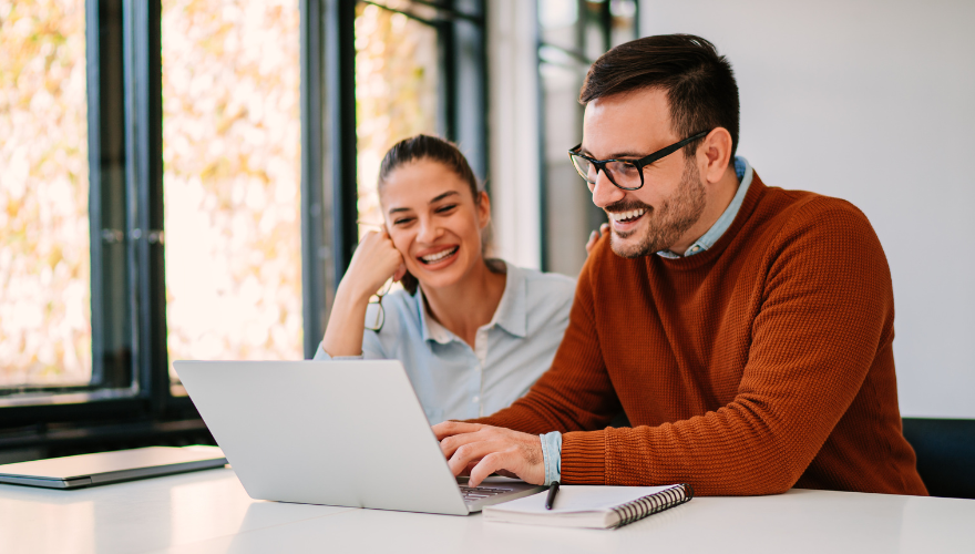 Man and woman at an office laughing while man is on laptop with notepad beside him